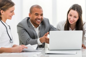 Two happy female doctors and African American businessman working together. They are using computer at doctor's office.